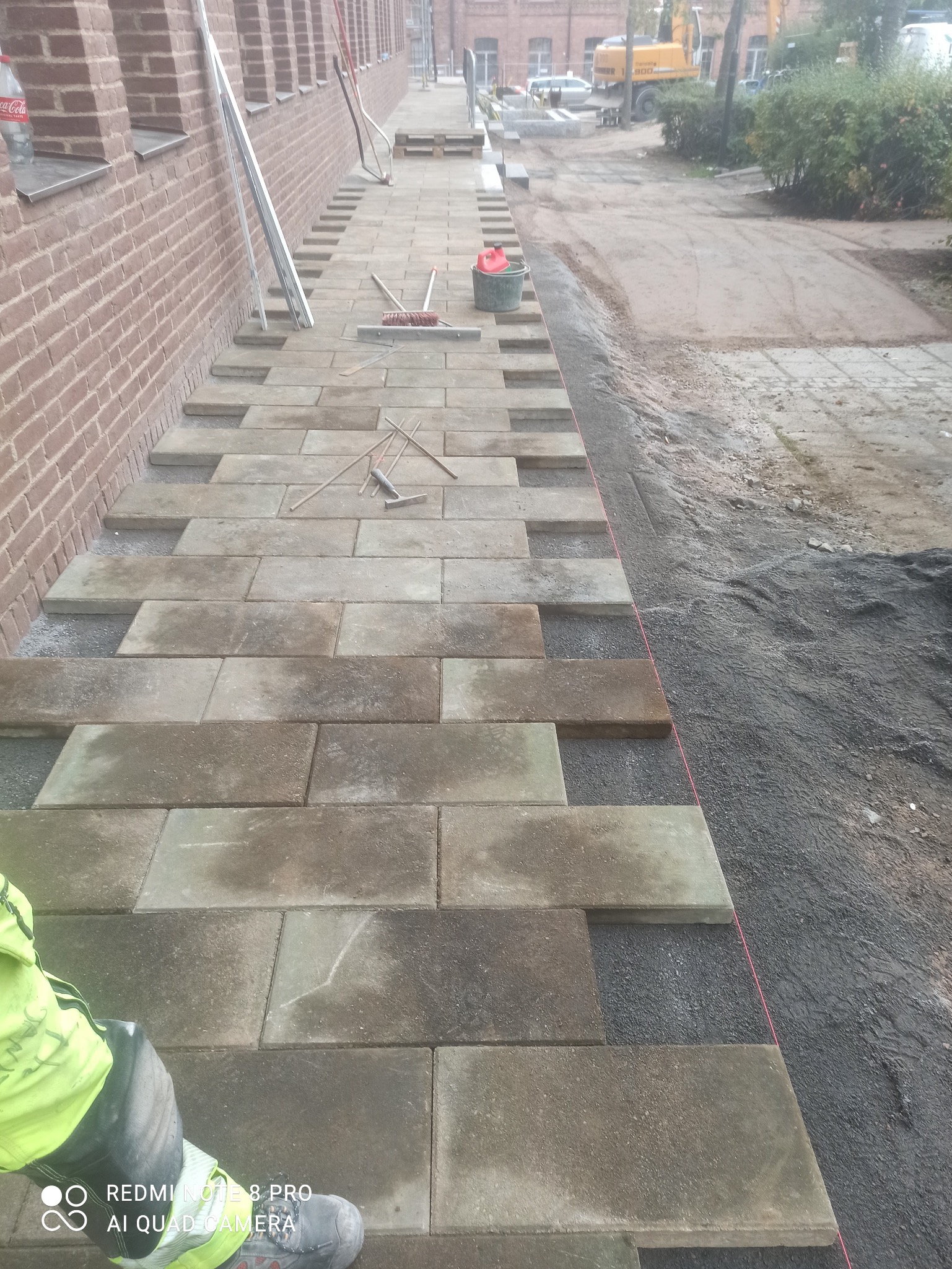 Fragment układanej ścieżki z kostki brukowej przy budynku z cegły. Widoczne narzędzia, piasek i częściowo ułożona nawierzchnia. W tle maszyna budowlana.