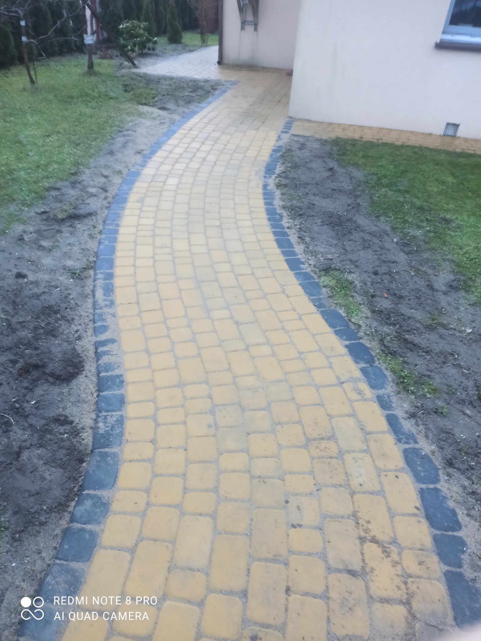 Żółta kostka brukowa ułożona w łukowaty chodnik z ciemnoszarą obwódką, prowadzący przez trawnik w kierunku budynku. Widok z góry, lekko zniekształcona perspektywa.