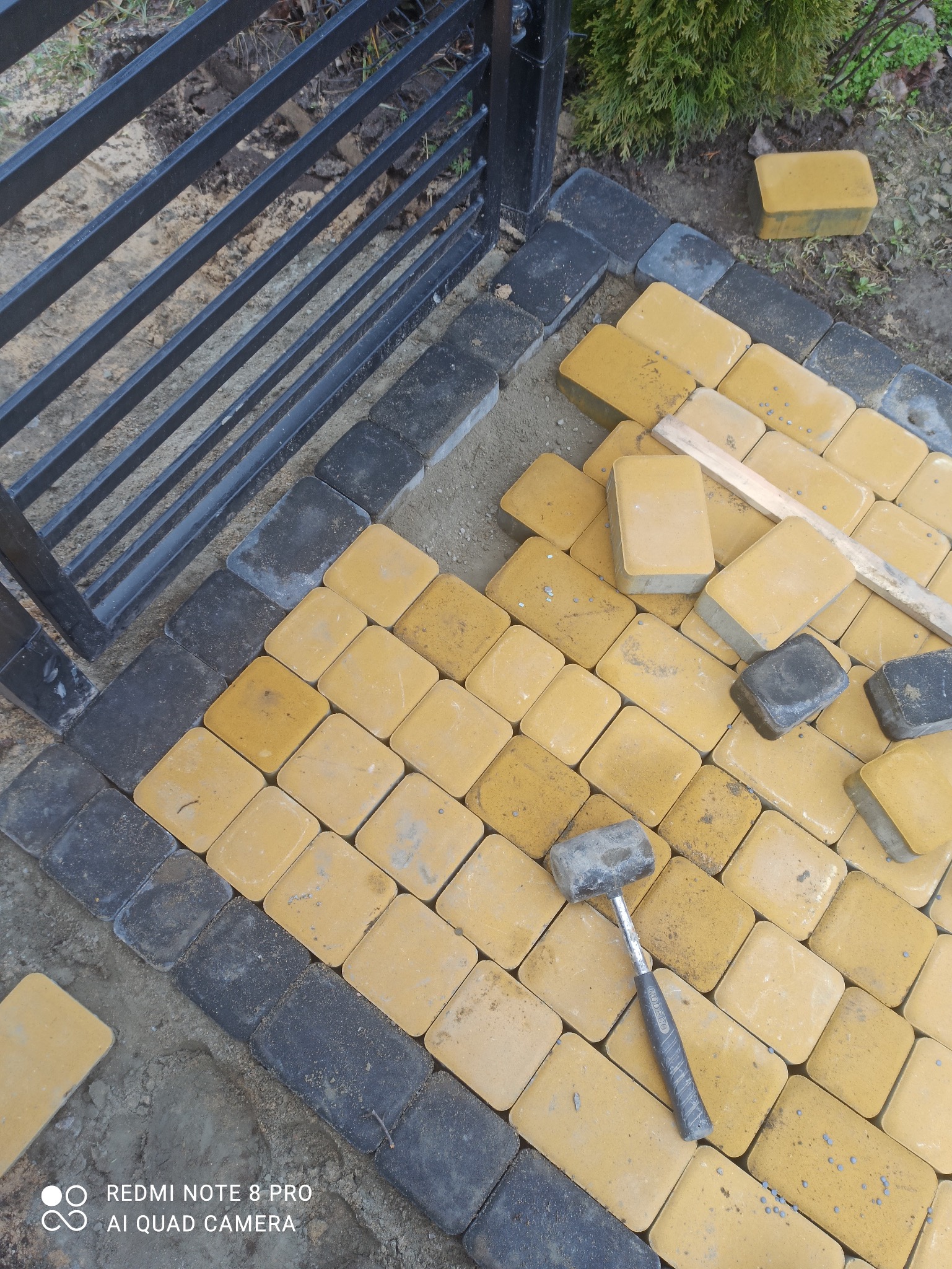 Żółto-czarna kostka brukowa ułożona przy metalowym ogrodzeniu, widoczny gumowy młotek i drewniana listwa. Częściowo zakończone prace brukarskie.