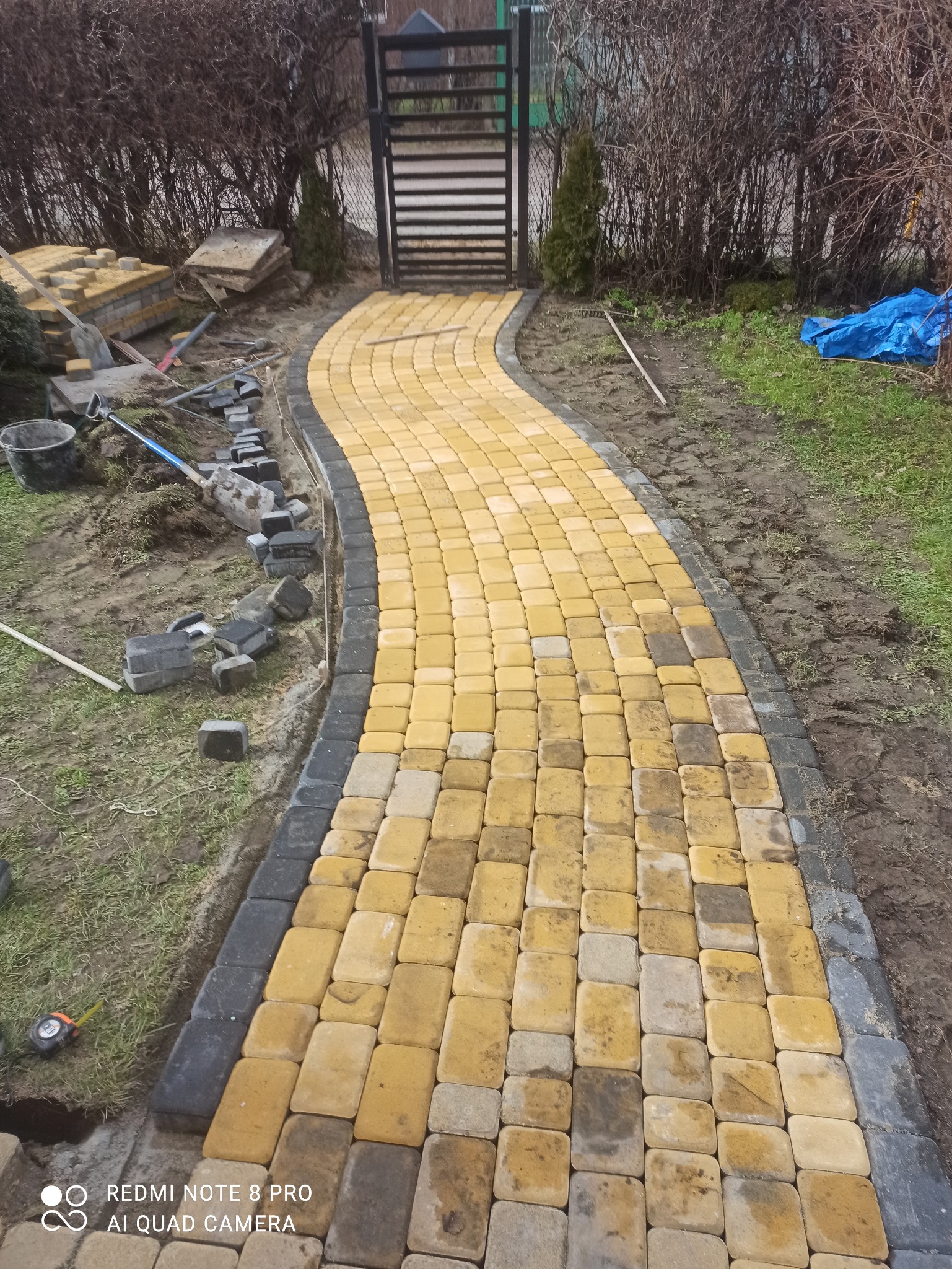 Żółto-szara ścieżka z kostki brukowej prowadząca do furtki, częściowo ułożona, z widocznymi narzędziami i stertą kostki obok. Widok z góry.