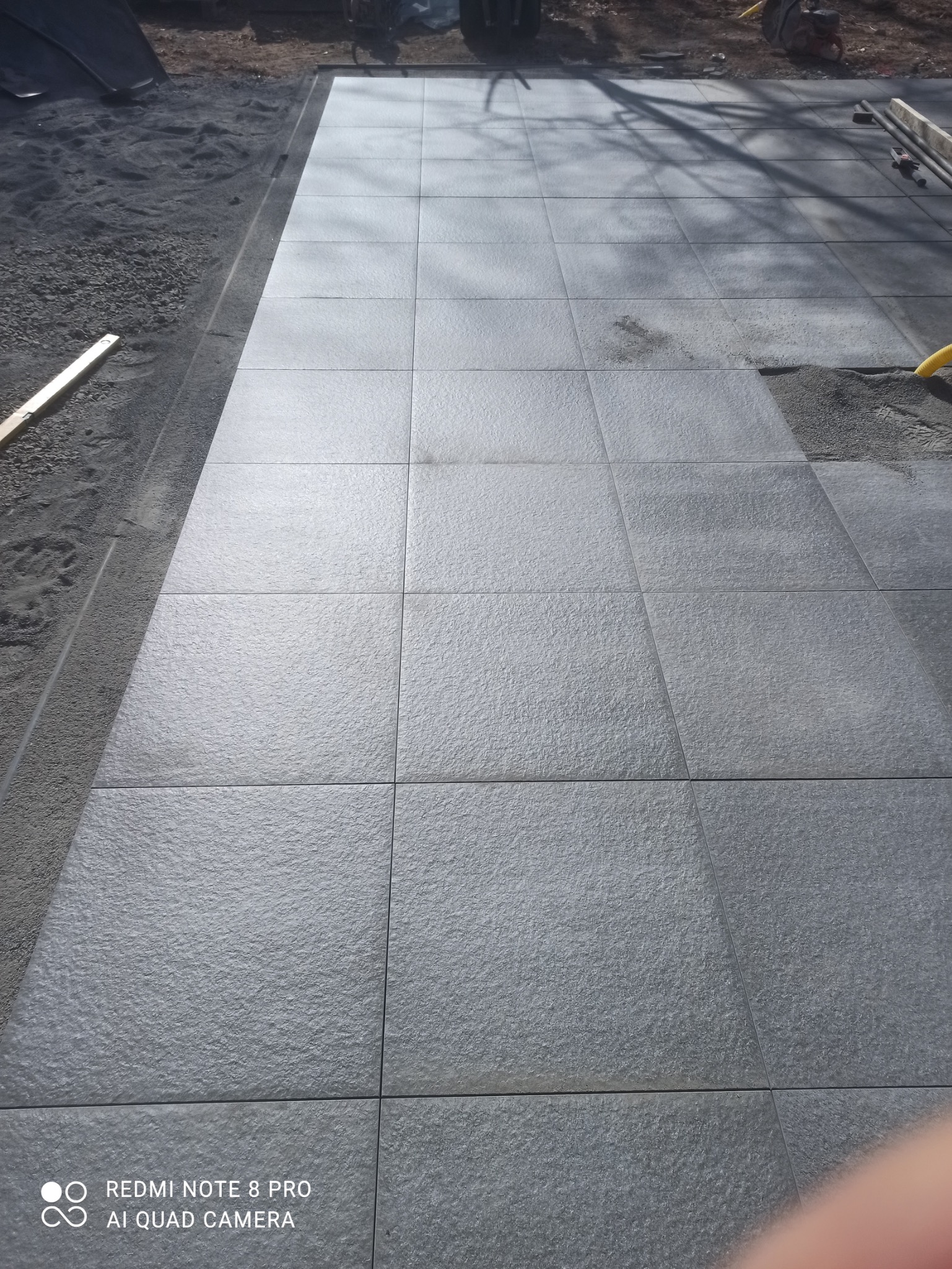 Ułożona, szara kostka brukowa na podwórku, widok z góry. Część powierzchni jeszcze nieukończona, widoczny piasek i narzędzia. Minimalistyczny styl.