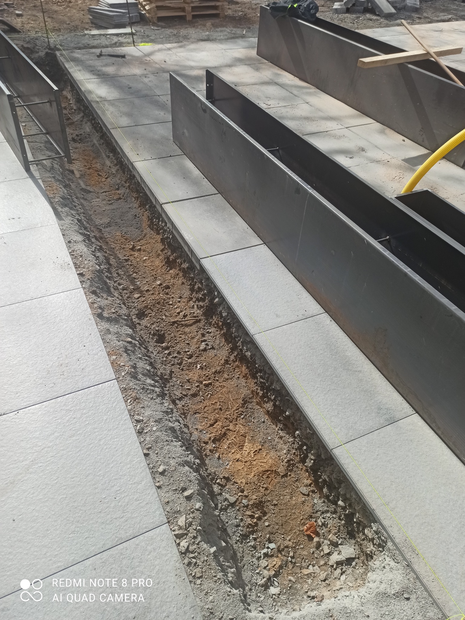 Przygotowanie podłoża pod kostkę brukową: wykop z widoczną warstwą ziemi, obrzeża z metalu i ułożone płyty. Żółta linka wyznacza linię. Etap prac.