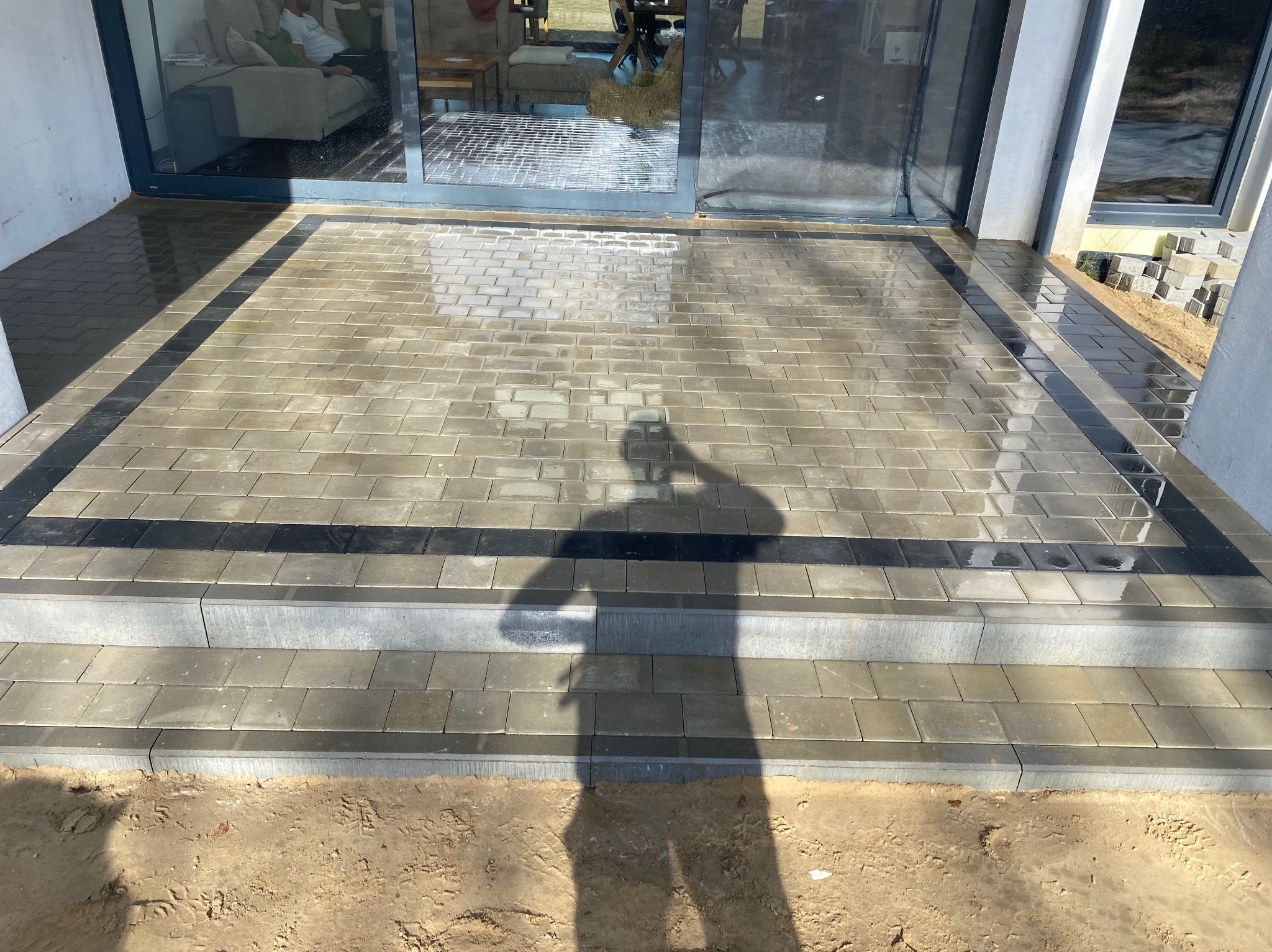 Ułożona kostka brukowa w odcieniach szarości z czarnym obramowaniem, tworząca schody wejściowe do domu z widocznym odbiciem w szklanych drzwiach.