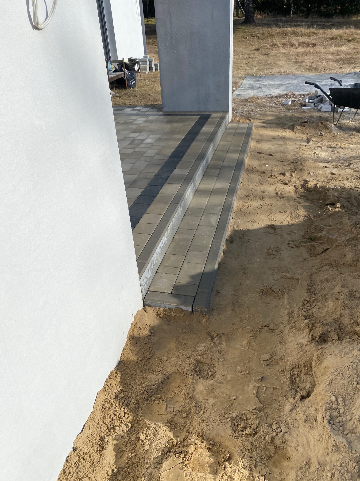 Schody z kostki brukowej w odcieniach szarości, prowadzące do budynku z jasną elewacją. Widoczny fragment terenu z piaskiem i narzędzia brukarskie w tle.