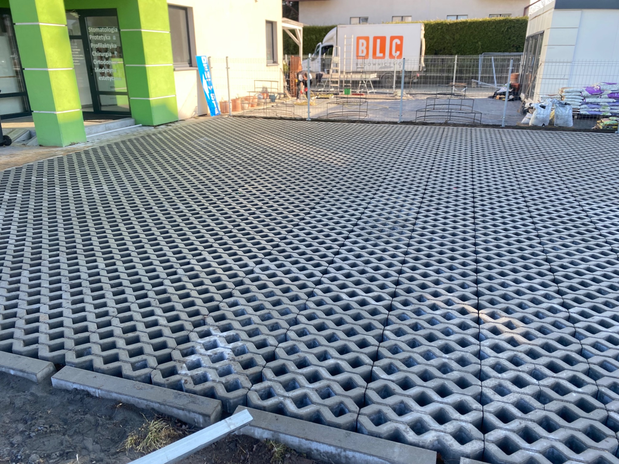Ułożona szara ażurowa kostka na podjeździe przed budynkiem z zielonymi kolumnami. W tle widoczny samochód dostawczy i ogrodzenie.