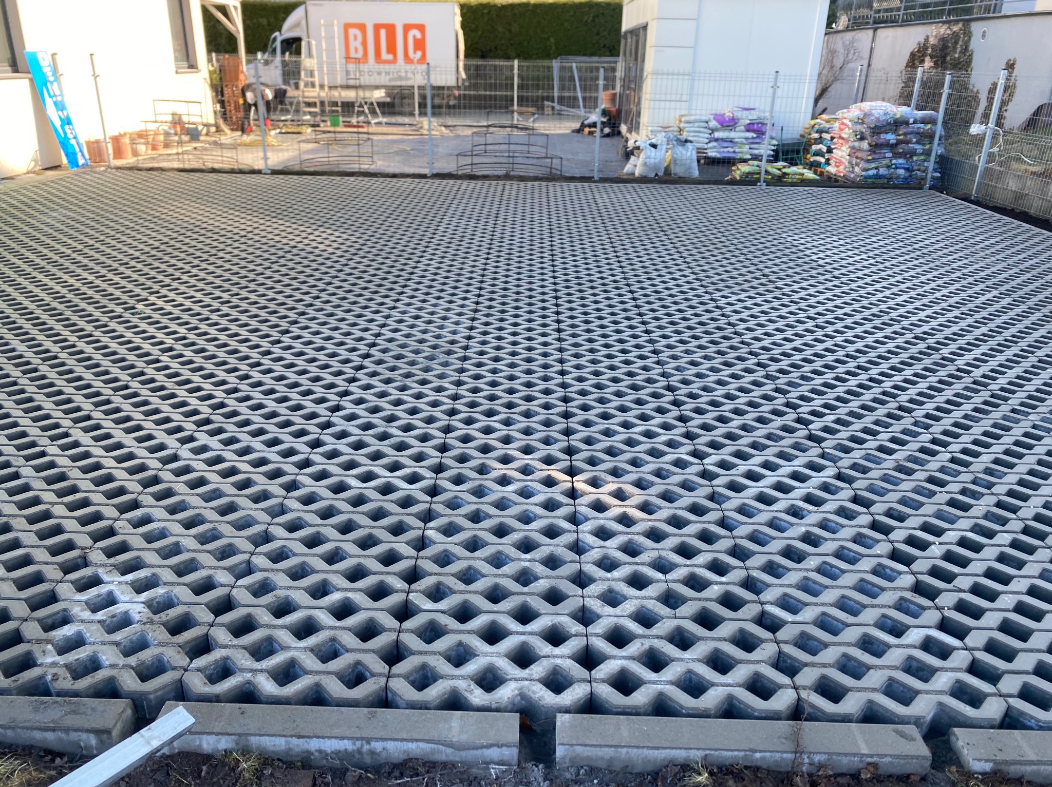 Ułożona szara ażurowa kostka brukowa na podjeździe, widok z góry. W tle budynek, samochód dostawczy i worki z materiałami budowlanymi.