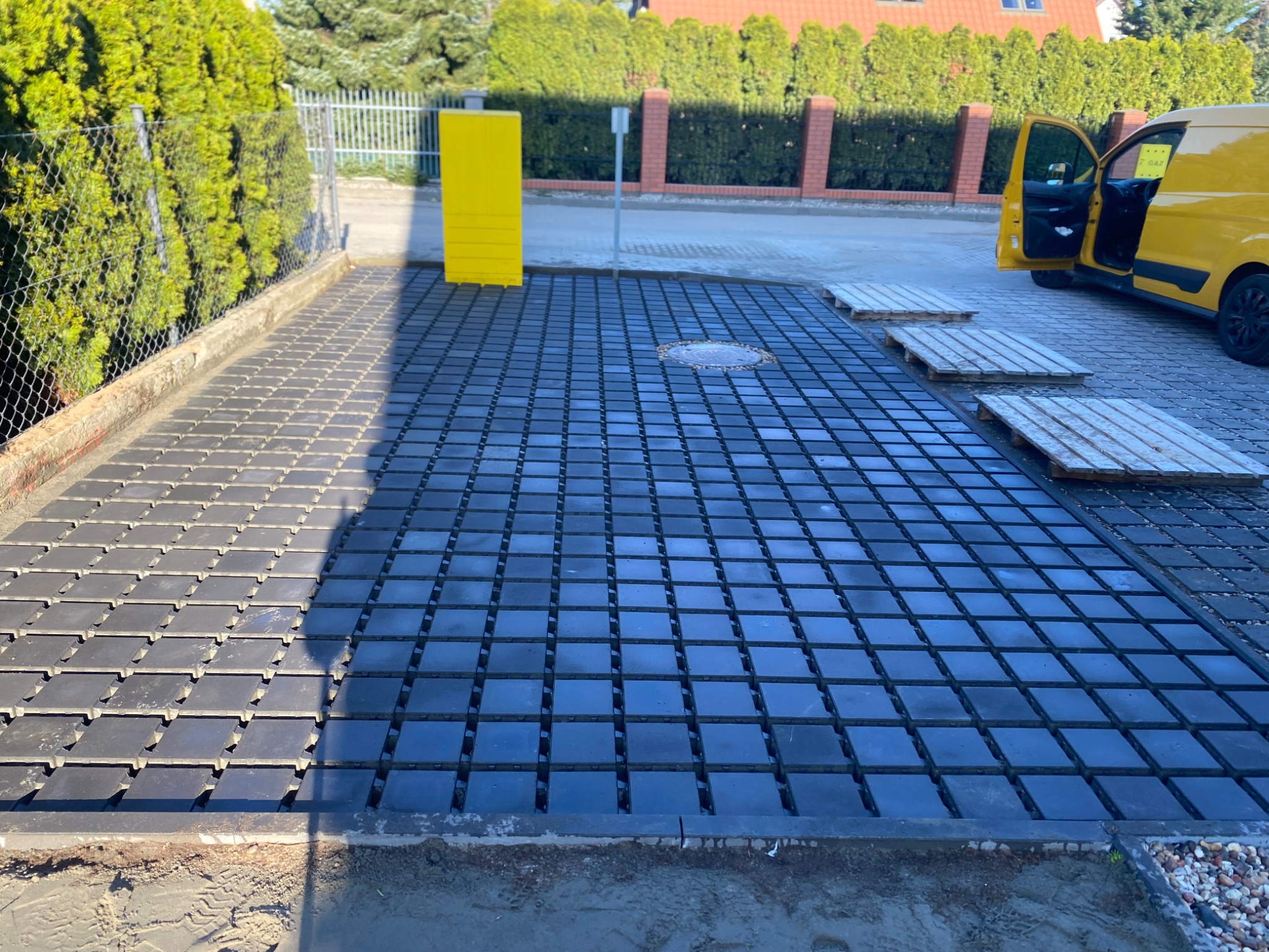 Częściowo ułożona, czarna kostka brukowa na podjeździe, z żółtym słupkiem i samochodem dostawczym w tle. Widoczne palety ułatwiające transport kostki.