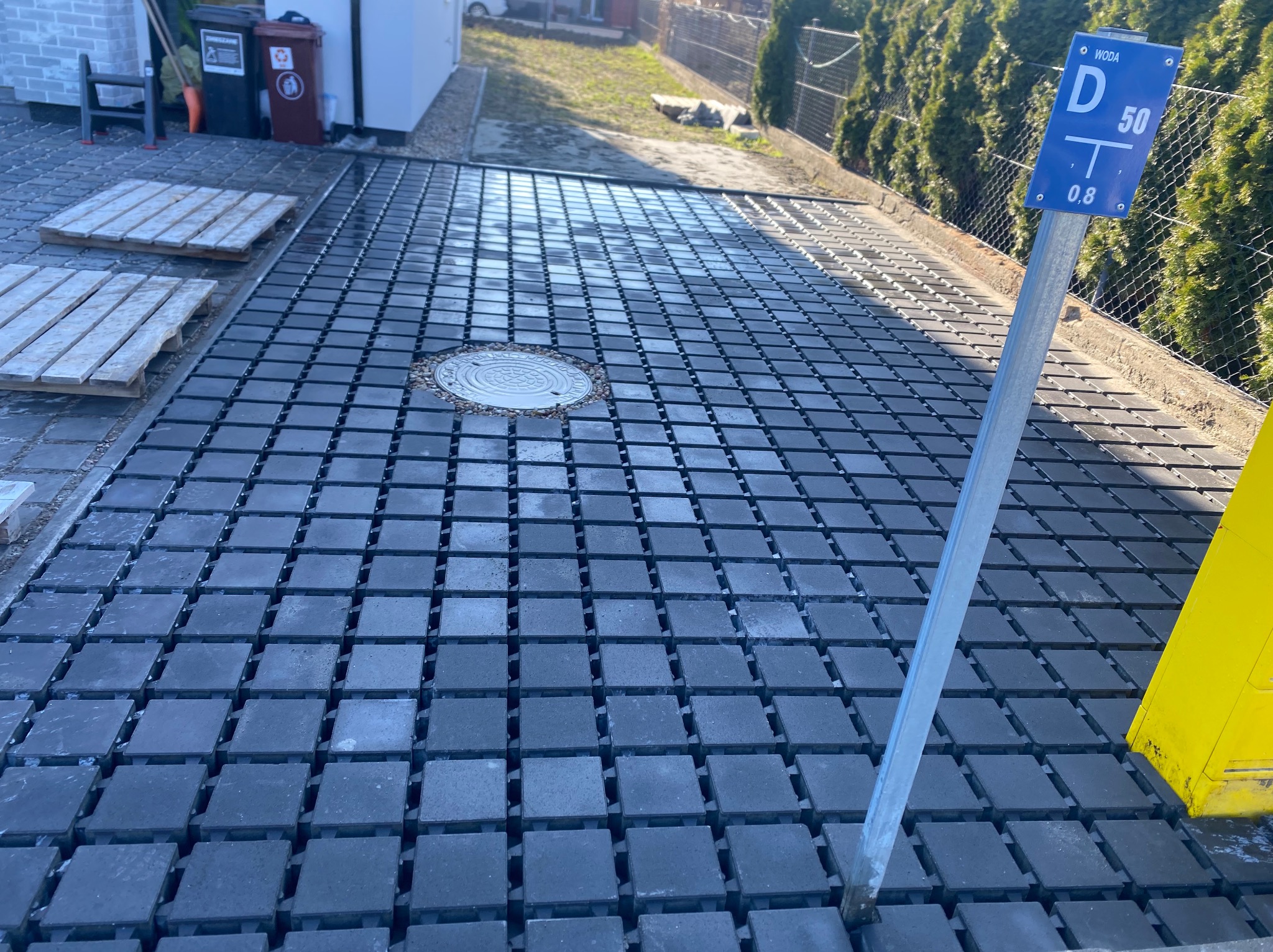 Ułożona, grafitowa kostka brukowa z kratką drenażową i studzienką kanalizacyjną. W tle zieleń i zabudowa. Widoczny słupek z oznaczeniem wodociągu.