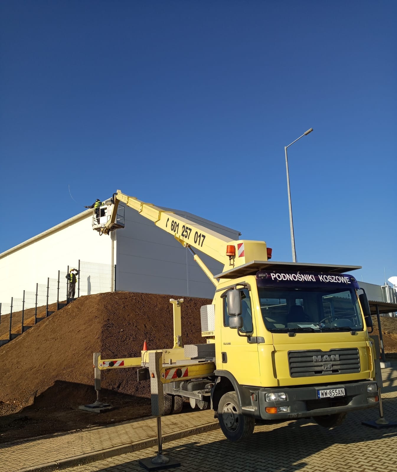 Żółty podnośnik koszowy MAN z dwoma pracownikami na wysokości, na tle białej hali i błękitnego nieba. Jeden z pracowników stoi na ziemi, drugi w koszu podnośnika.