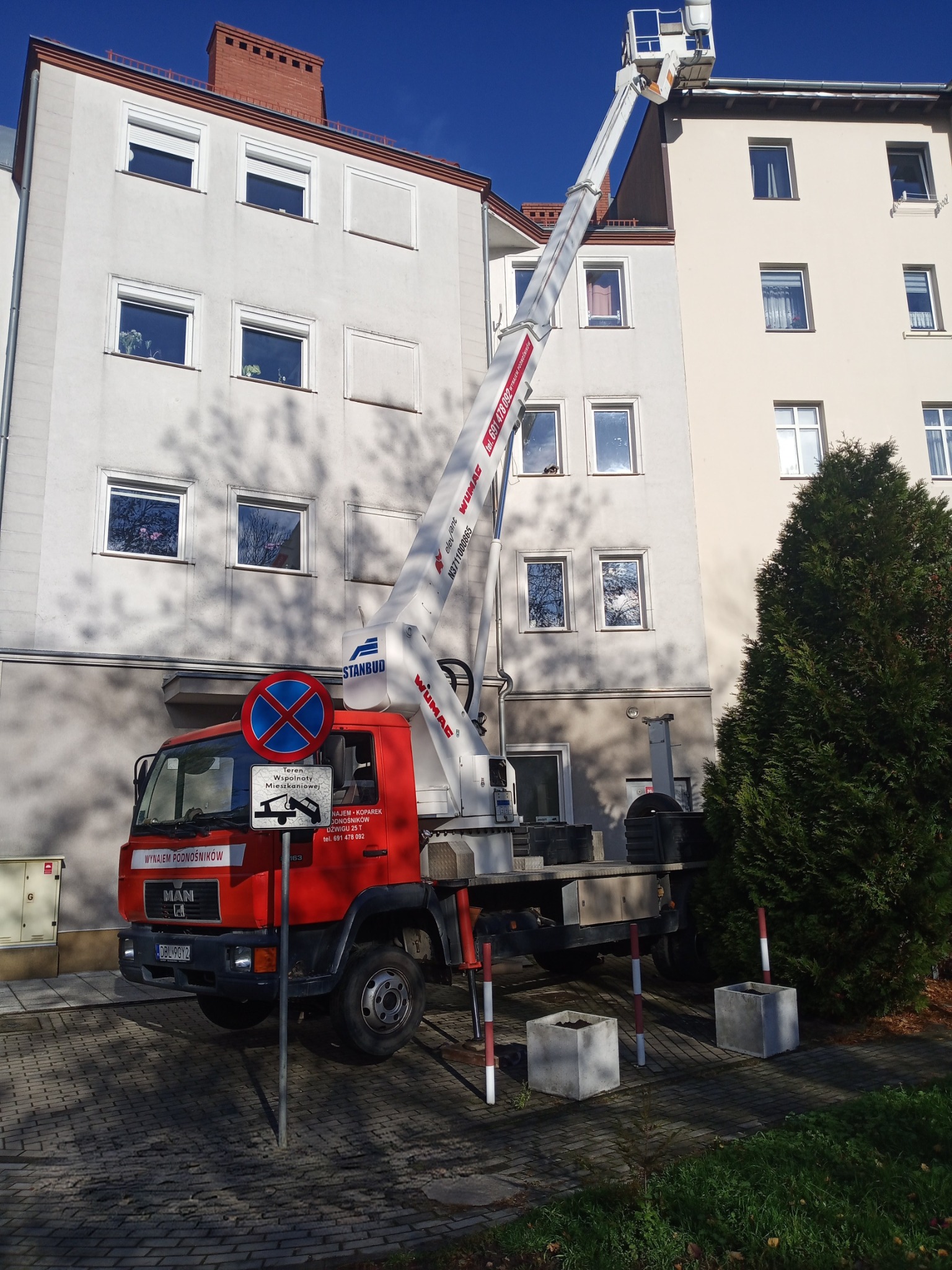 Czerwony podnośnik koszowy Stanbud na tle budynku mieszkalnego, z wysuniętym ramieniem i koszem przy rynnie. Widoczny znak zakazu postoju.