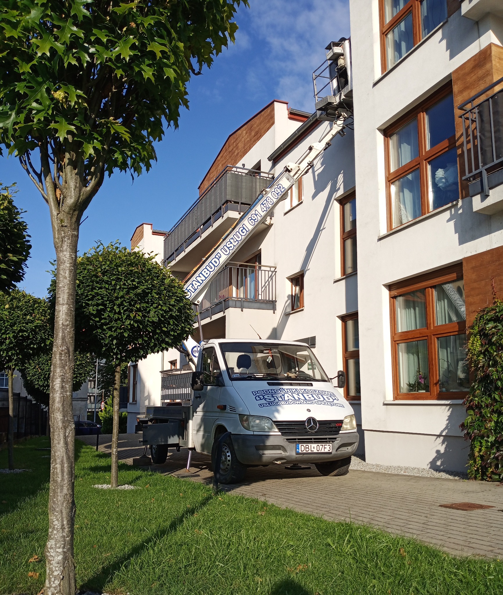 Podnośnik koszowy na tle budynku z balkonami, z wysuniętym ramieniem do okna. Widoczne drzewa i trawnik. Jasne, słoneczne oświetlenie.