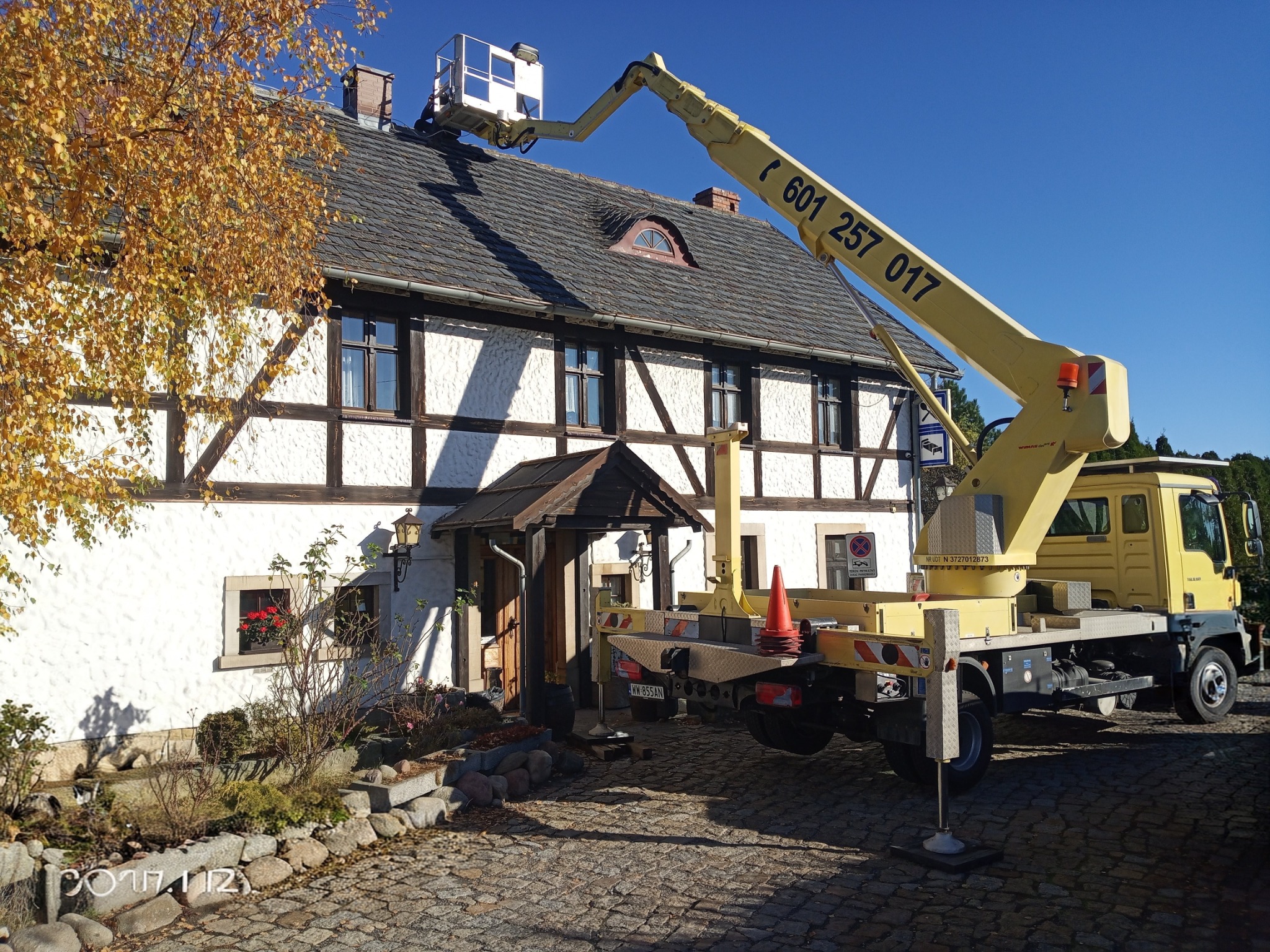 Żółty podnośnik koszowy na tle białego domu z muru pruskiego i brukowanej drogi. Kosz podnośnika przy dachu. Błękitne niebo w tle.