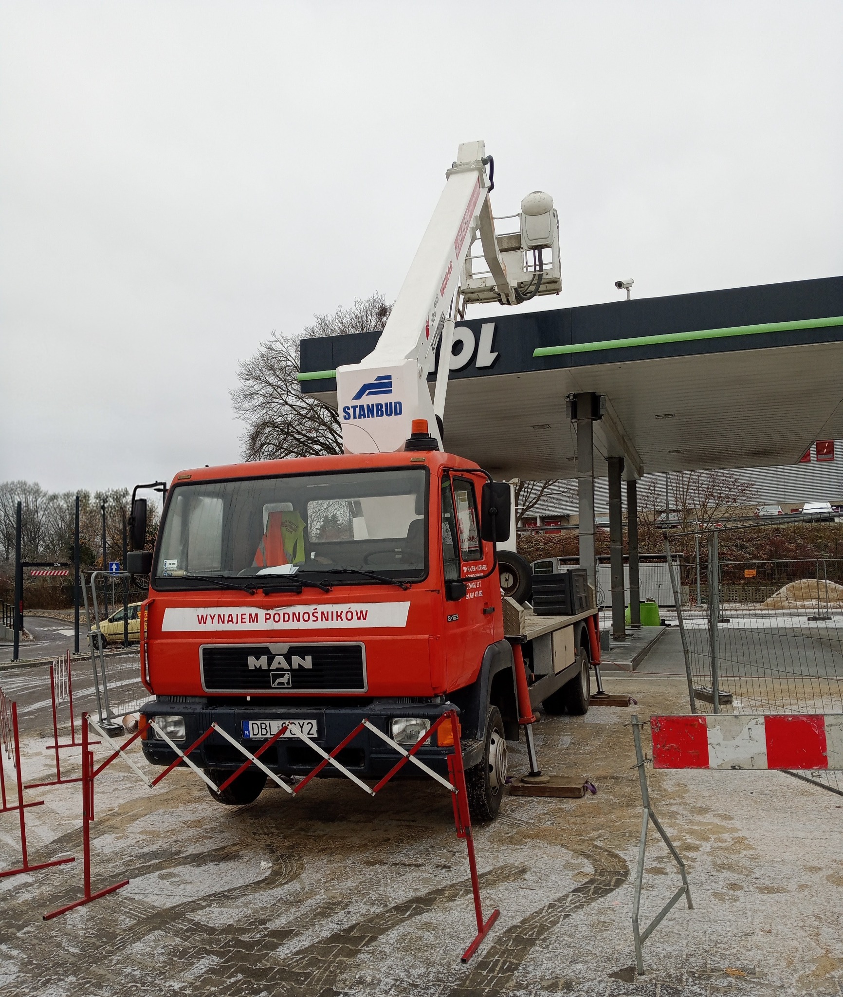 Pomarańczowy podnośnik koszowy MAN firmy Stanbud na tle stacji benzynowej, z wysięgnikiem skierowanym ku górze. Zabezpieczenia na ziemi.
