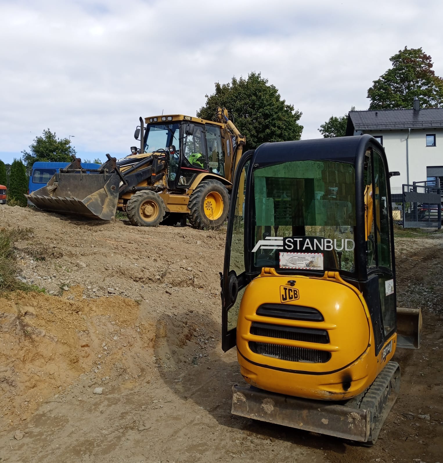 Żółta minikoparka JCB z otwartą kabiną na placu budowy, w tle koparko-ładowarka CAT i budynek mieszkalny. Na szybie naklejka z nazwą firmy.
