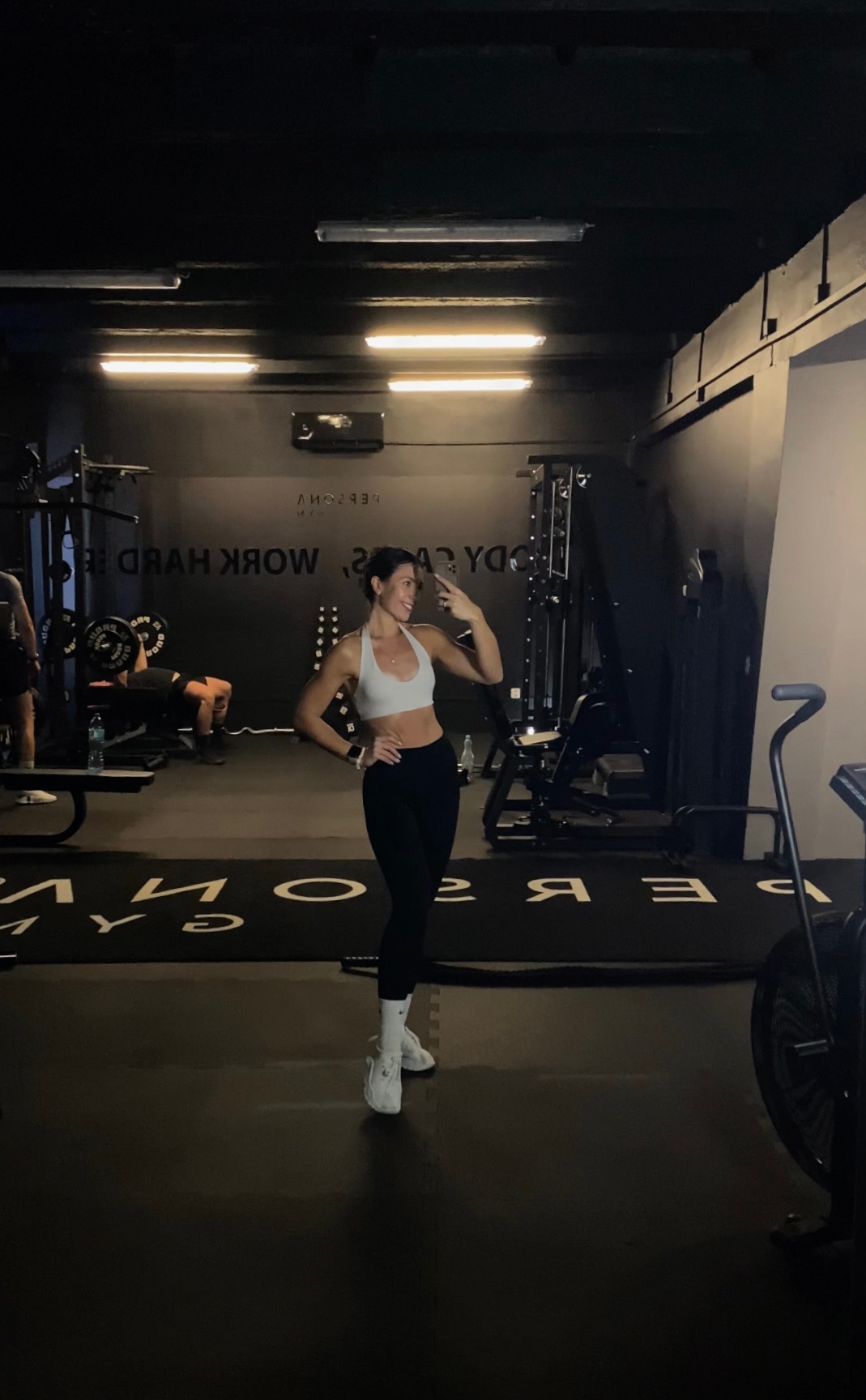 Uśmiechnięta kobieta w sportowym stroju robi selfie w siłowni. W tle sprzęt do ćwiczeń i osoba podnosząca ciężary. Ciemne wnętrze, jasne oświetlenie.