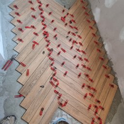 Krzysztof Wiśniewski - Układanie parkietu w jodełkę z użyciem czerwonych klipsów poziomujących. Widoczny kubeł z klinami i fragment buta wykonawcy. Prace w toku, surowe ściany.