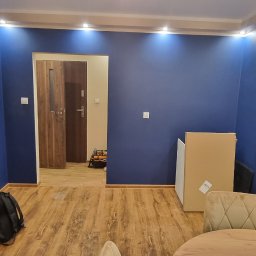 Krzysztof Wiśniewski - Pokój po malowaniu na intensywny, ciemny odcień niebieskiego. Widoczne wykończone ściany, drewniana podłoga i fragmenty mebli. Oświetlenie sufitowe punktowe.