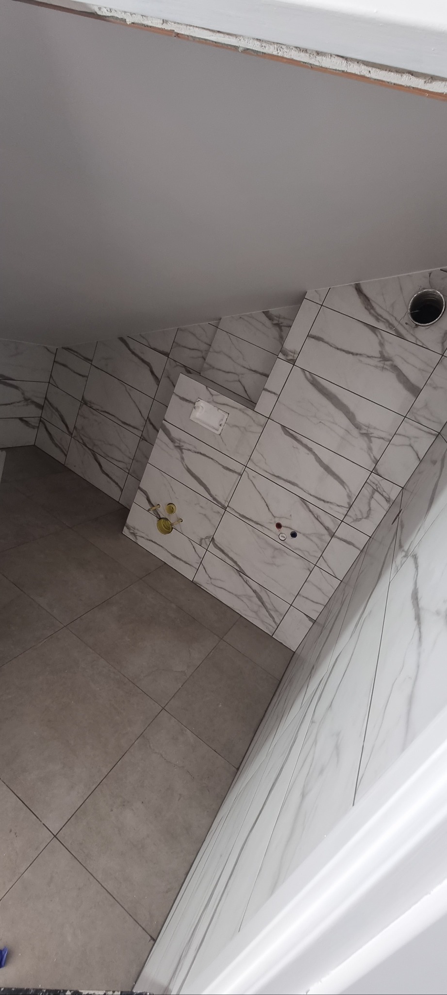 Wykończona łazienka z marmurowymi płytkami na ścianach i szarą podłogą. Widoczne przyłącza wodne i wentylacja. Ujęcie z góry, podkreślające geometrię pomieszczenia.