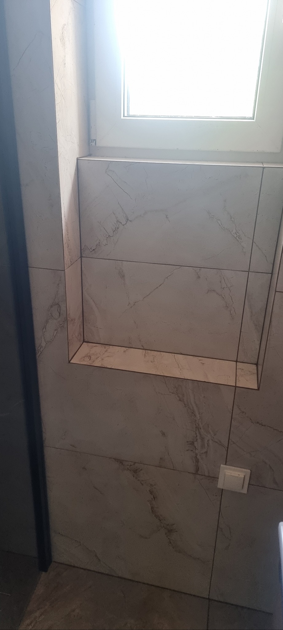 Wykończona wnęka w łazience z płytkami imitującymi marmur, oświetlona naturalnym światłem z okna. Widoczny fragment włącznika światła i ramy drzwi.