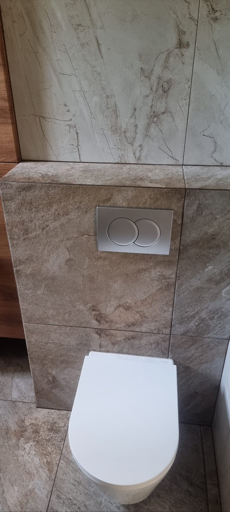 Biała toaleta wisząca z chromowanym przyciskiem spłukującym na tle beżowych płytek imitujących marmur. Widoczny fragment zabudowy z płytek i podłogi.