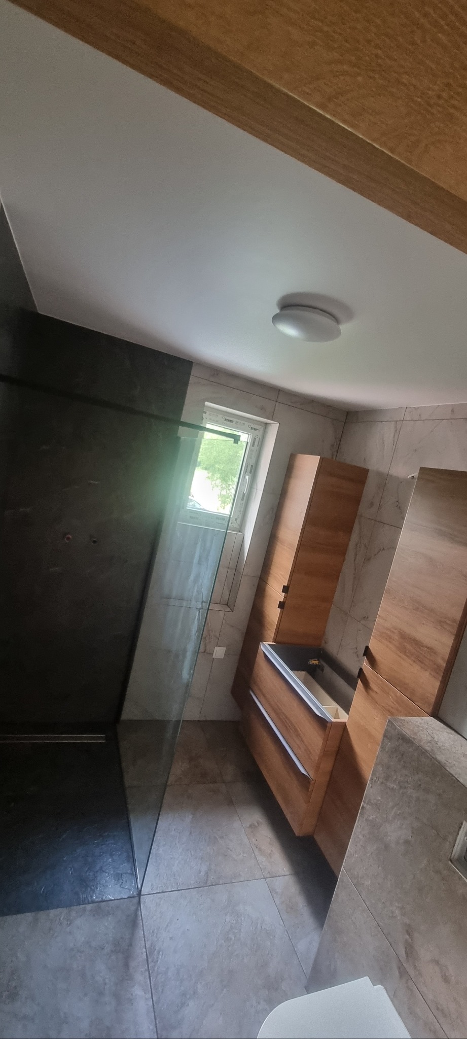 Nowoczesna łazienka z kabiną prysznicową typu walk-in, drewnianą szafką pod umywalką i marmurowymi płytkami. Widoczne okno i oświetlenie sufitowe.