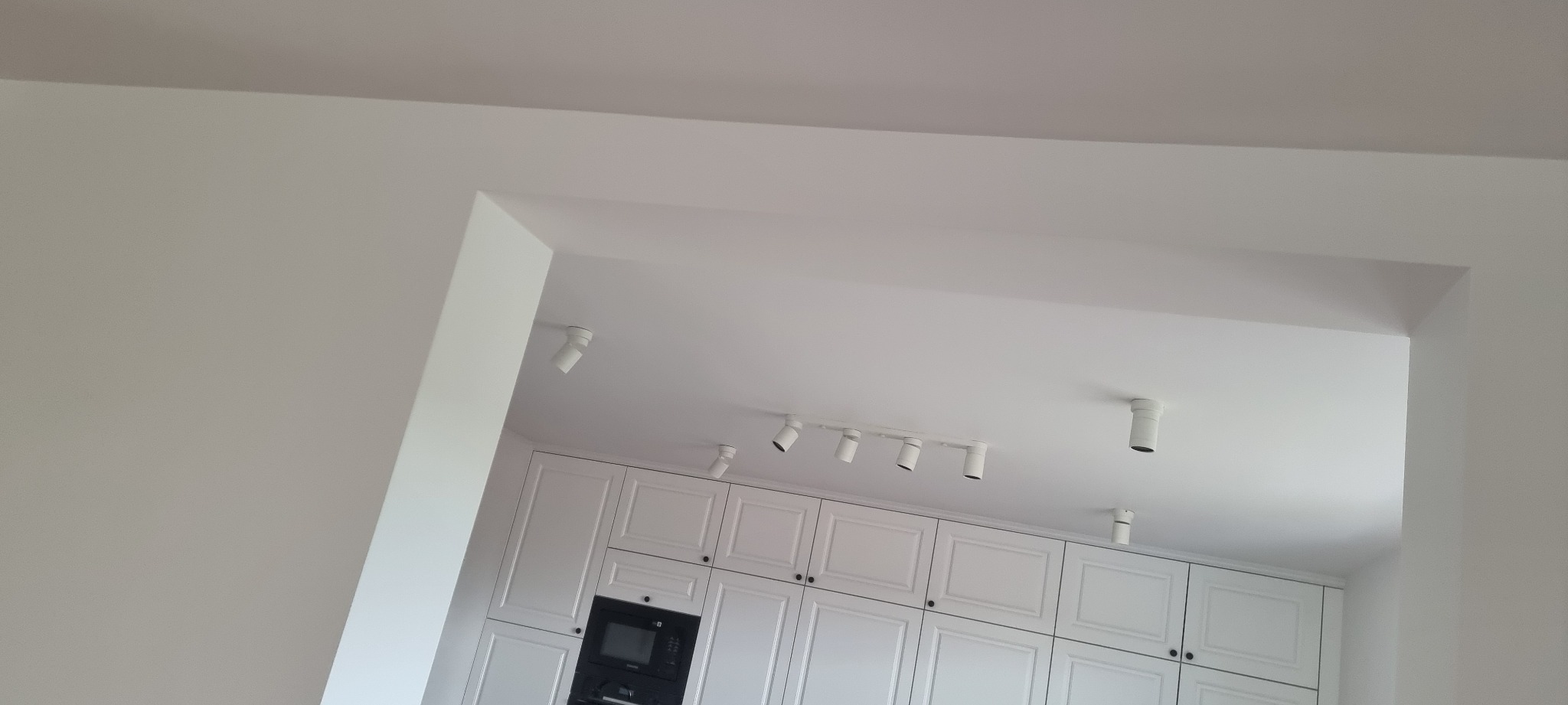 Biała kuchnia z oświetleniem punktowym na suficie, widok z dołu przez otwór w ścianie. Minimalistyczny design, jasne wnętrze.