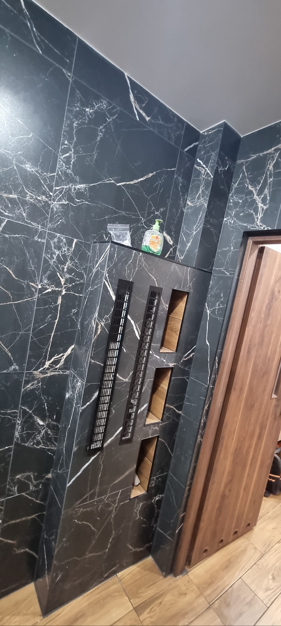 Wykończona łazienka z czarnymi płytkami imitującymi marmur, z wnękami i kratkami wentylacyjnymi. Widoczna butelka z mydłem i drewniane drzwi.