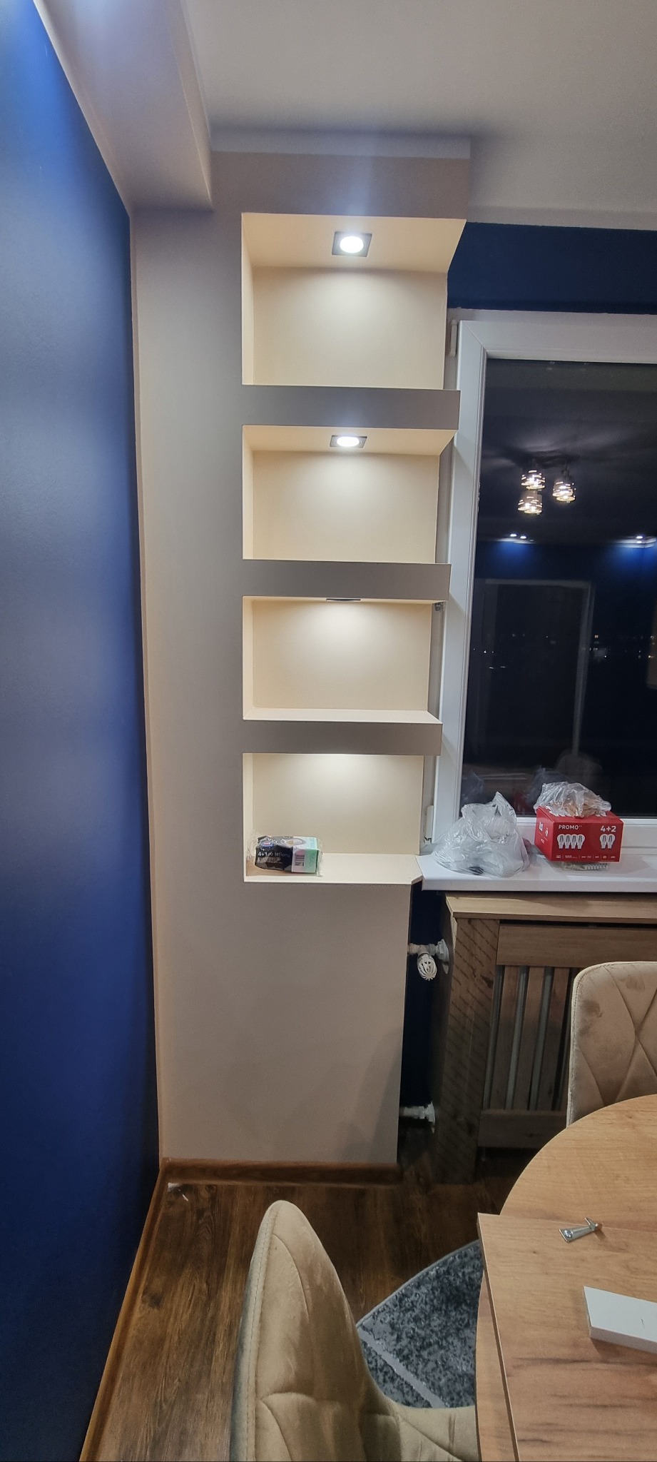 Narożna ściana z wnękami oświetlonymi punktowo, pomalowana na beżowo, kontrastująca z niebieską ścianą obok okna. Widoczne elementy wyposażenia wnętrza.