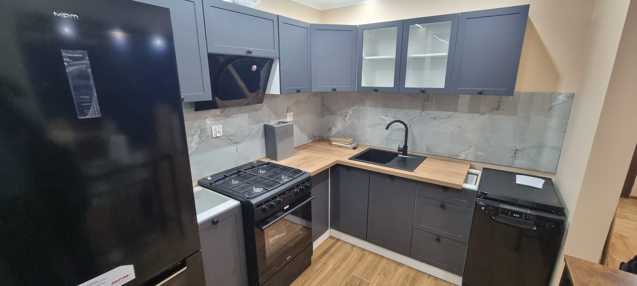 Nowoczesna kuchnia w odcieniach szarości z drewnianym blatem, czarnym zlewem i sprzętem AGD. Widoczne szafki z przeszkleniami i marmurowe wykończenie ściany.