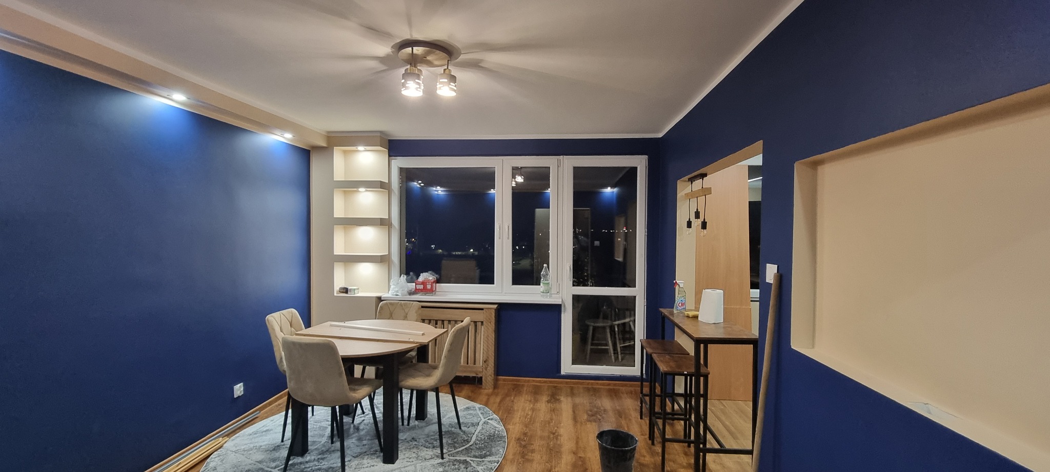 Nowoczesne wnętrze: granatowe ściany, drewniana podłoga, stół z krzesłami i barek. Oświetlenie sufitowe i wnęki ścienne. Widok na nocne miasto przez okno.