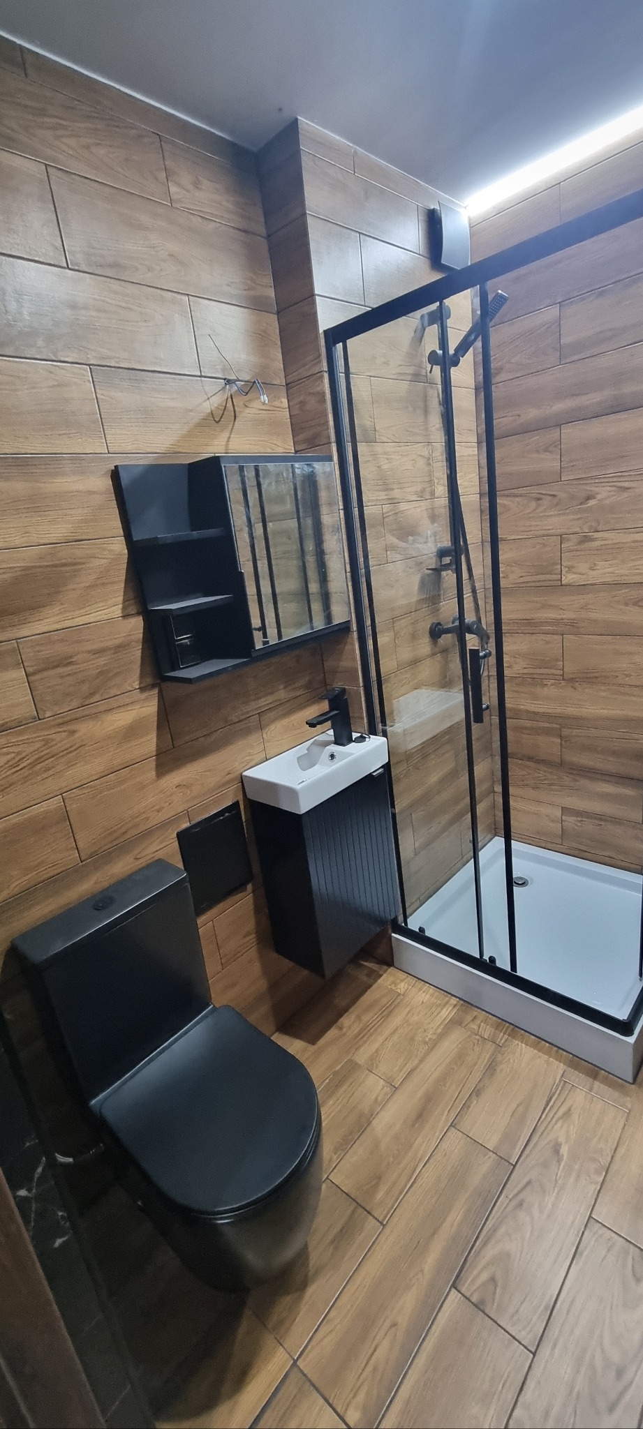 Nowoczesna łazienka z drewnopodobnymi płytkami, czarną armaturą i kabiną prysznicową typu walk-in. Umywalka z szafką i lustro z półkami w minimalistycznym stylu.