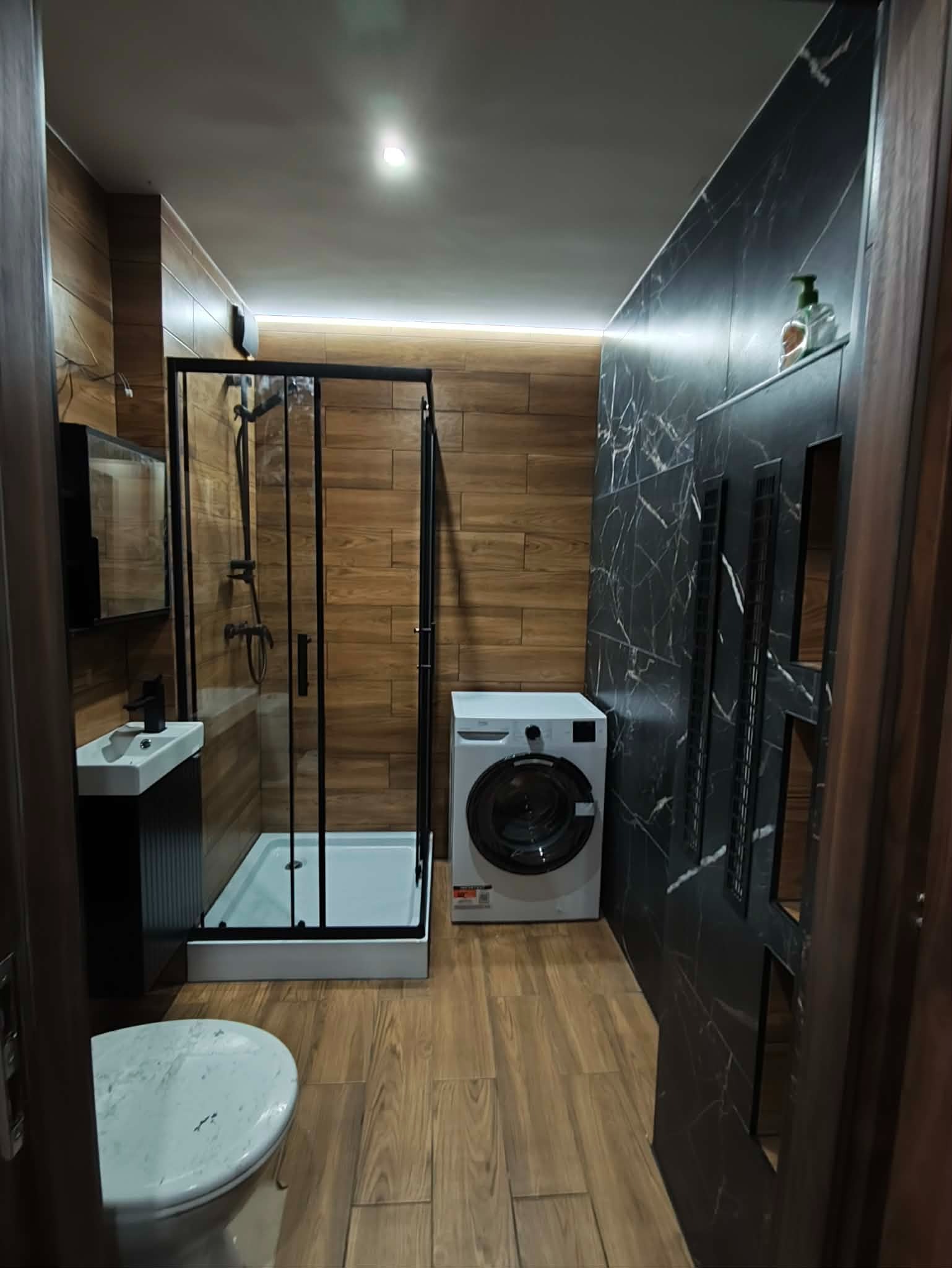 Nowoczesna łazienka z kabiną prysznicową typu walk-in, pralką i umywalką na szafce. Ściany wyłożone płytkami imitującymi drewno i czarny marmur.