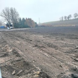 Masterkop - Przygotowany, wyrównany teren pod budowę z widocznymi śladami opon, w tle budynek i drzewa. Pochmurna pogoda, ziemia ciemna, świeżo obrobiona.