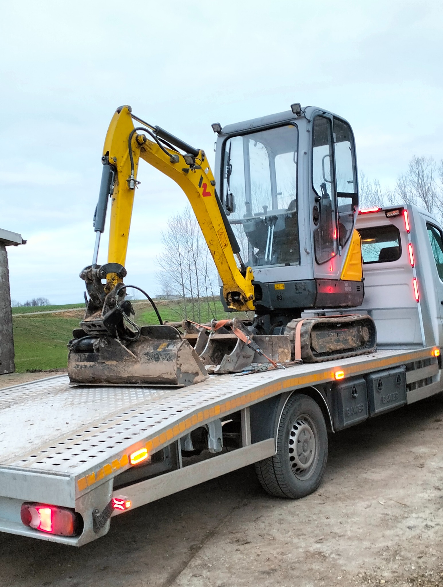 Żółto-szara mini koparka na platformie lawety, zabezpieczona pasami. Widoczne gąsienice, łyżka koparki i kabina. W tle zielone wzgórze i nagie drzewa.