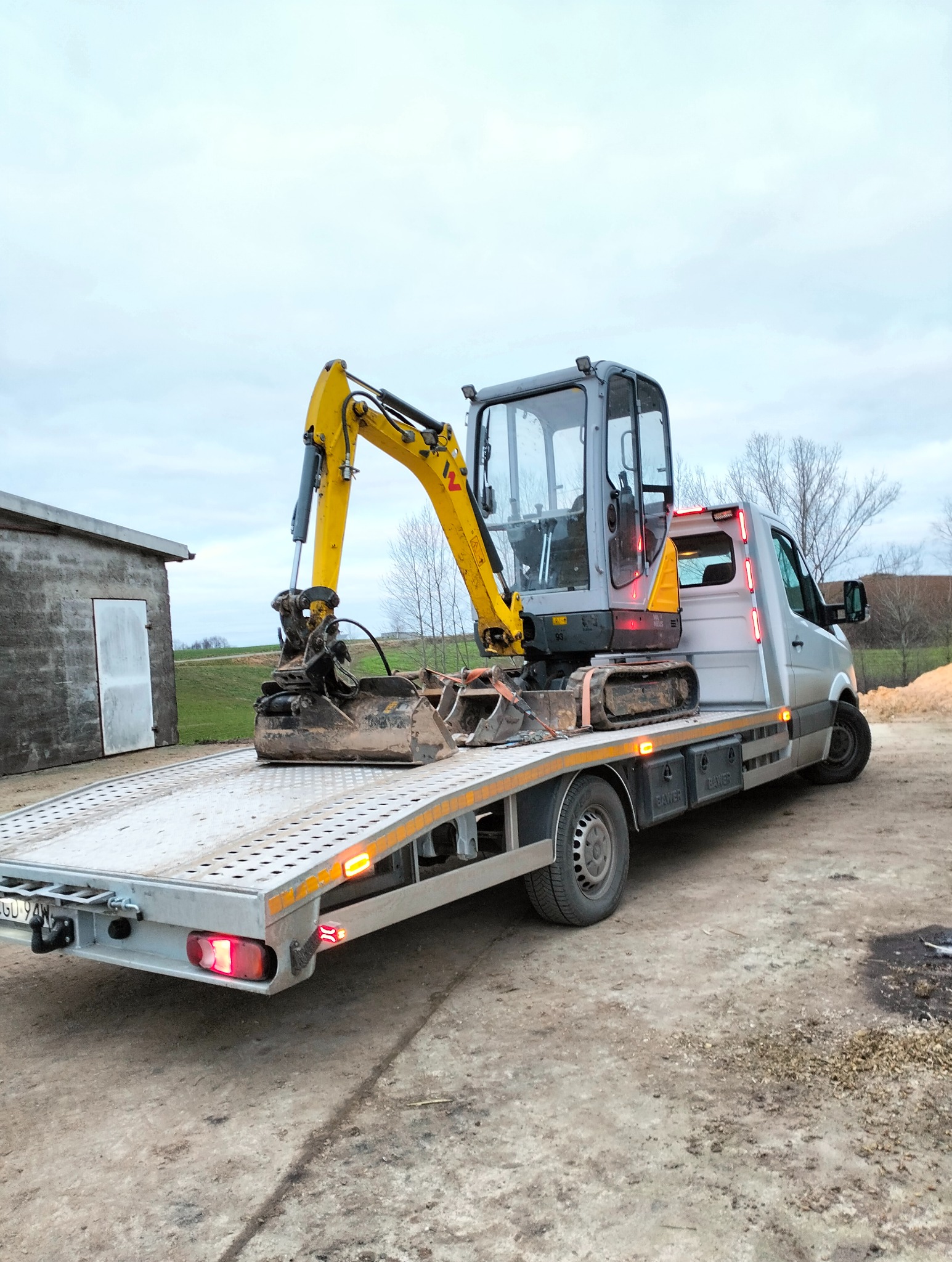 Żółto-szara mini koparka na lawecie srebrnego samochodu transportowego. W tle budynek gospodarczy i pole. Sprzęt budowlany gotowy do transportu na miejsce robót.