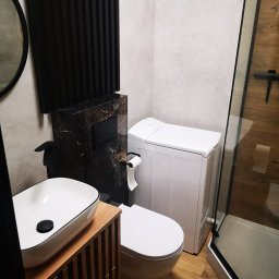Glazuro DOLNYŚLĄSK - Nowoczesna łazienka z drewnianą podłogą i szafką pod umywalkę, marmurową obudową WC, białą pralką i kabiną prysznicową z drewnianymi panelami.