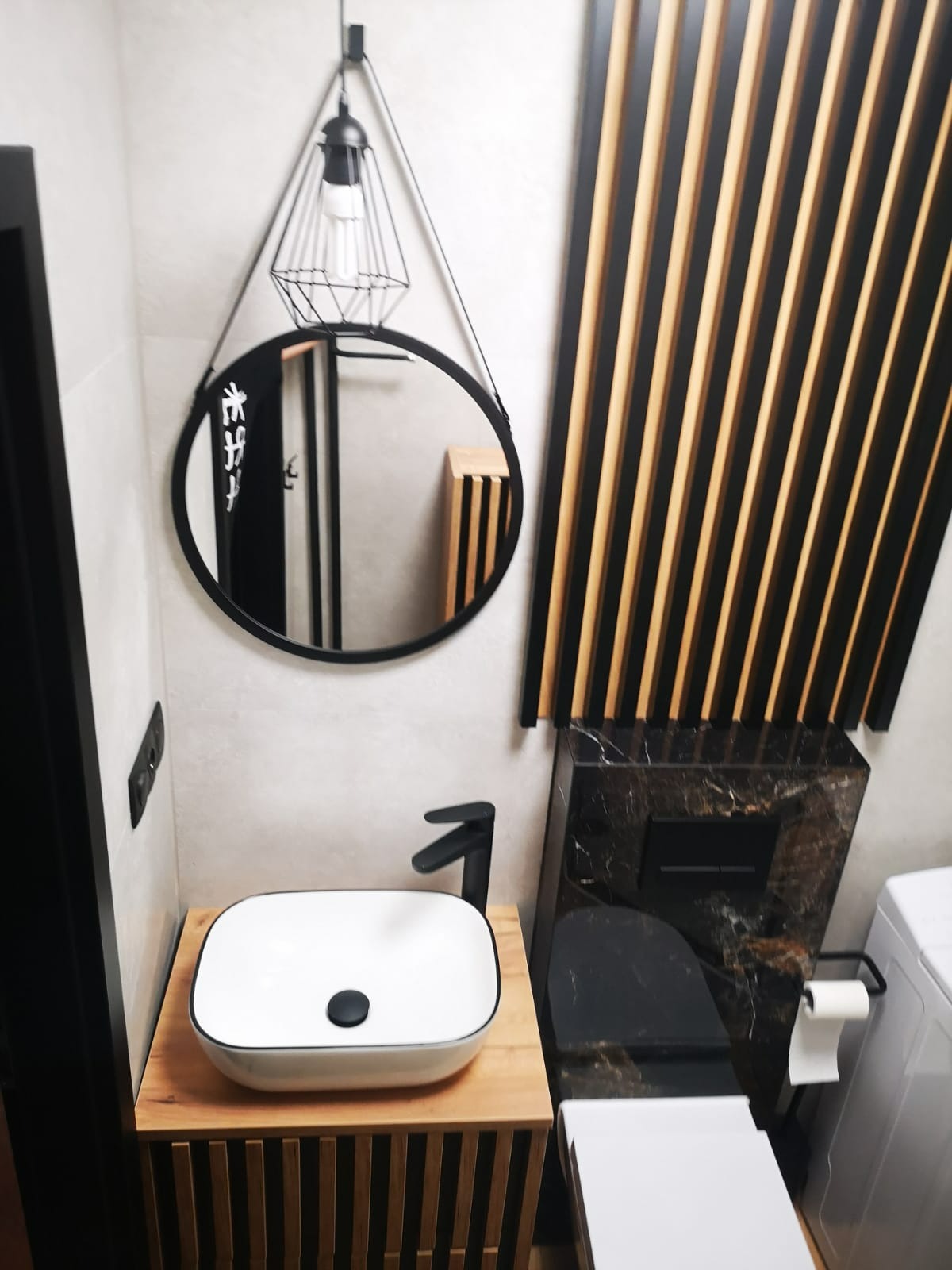 Nowoczesna łazienka z okrągłym lustrem, wiszącą lampą i umywalką na drewnianej szafce. Ściana zdobiona drewnianymi lamelami. Marmurowa obudowa toalety.