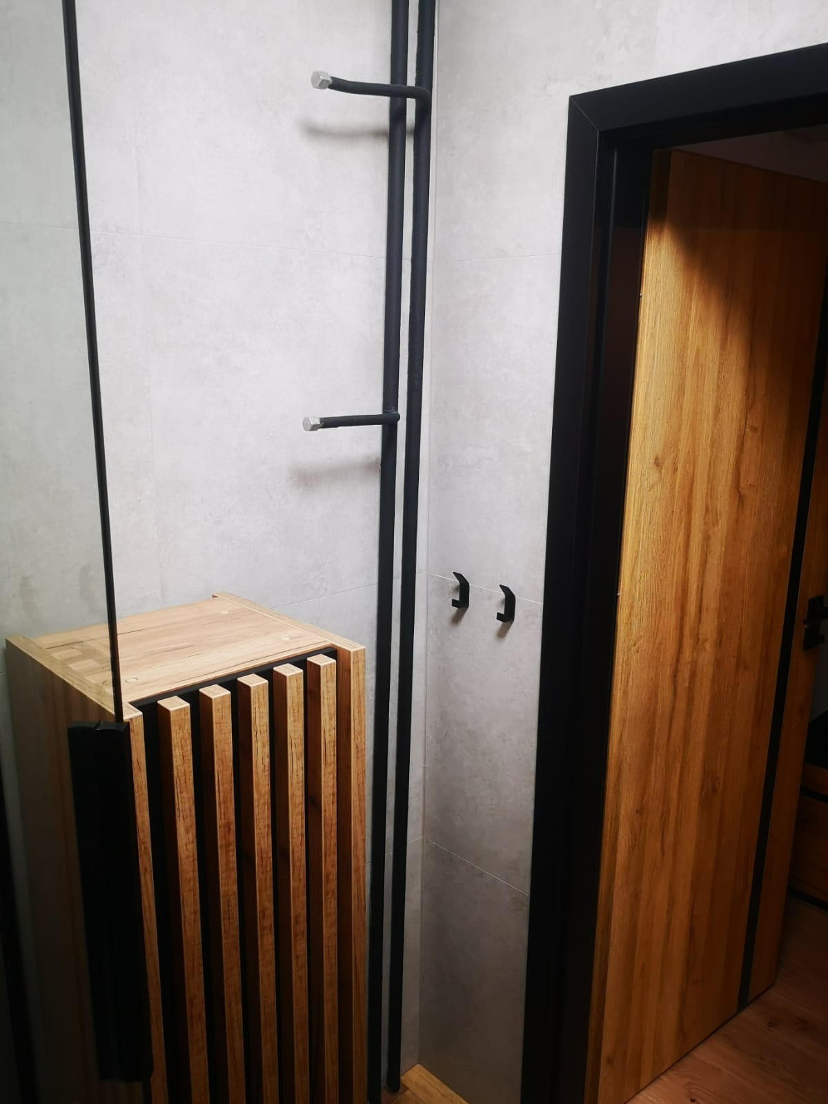 Nowoczesna łazienka z drewnianą szafką pod umywalkę z lamelami, czarnymi akcentami i szklaną kabiną prysznicową. Minimalistyczny design, jasne płytki.