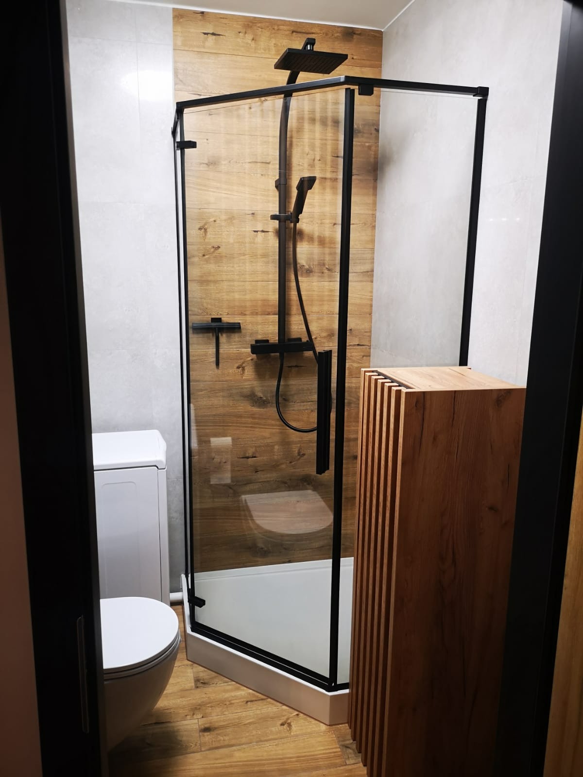 Nowoczesna kabina prysznicowa z czarnymi profilami i deszczownicą, drewnopodobne płytki na ścianie, drewniany słupek dekoracyjny, toaleta i pralka w tle.