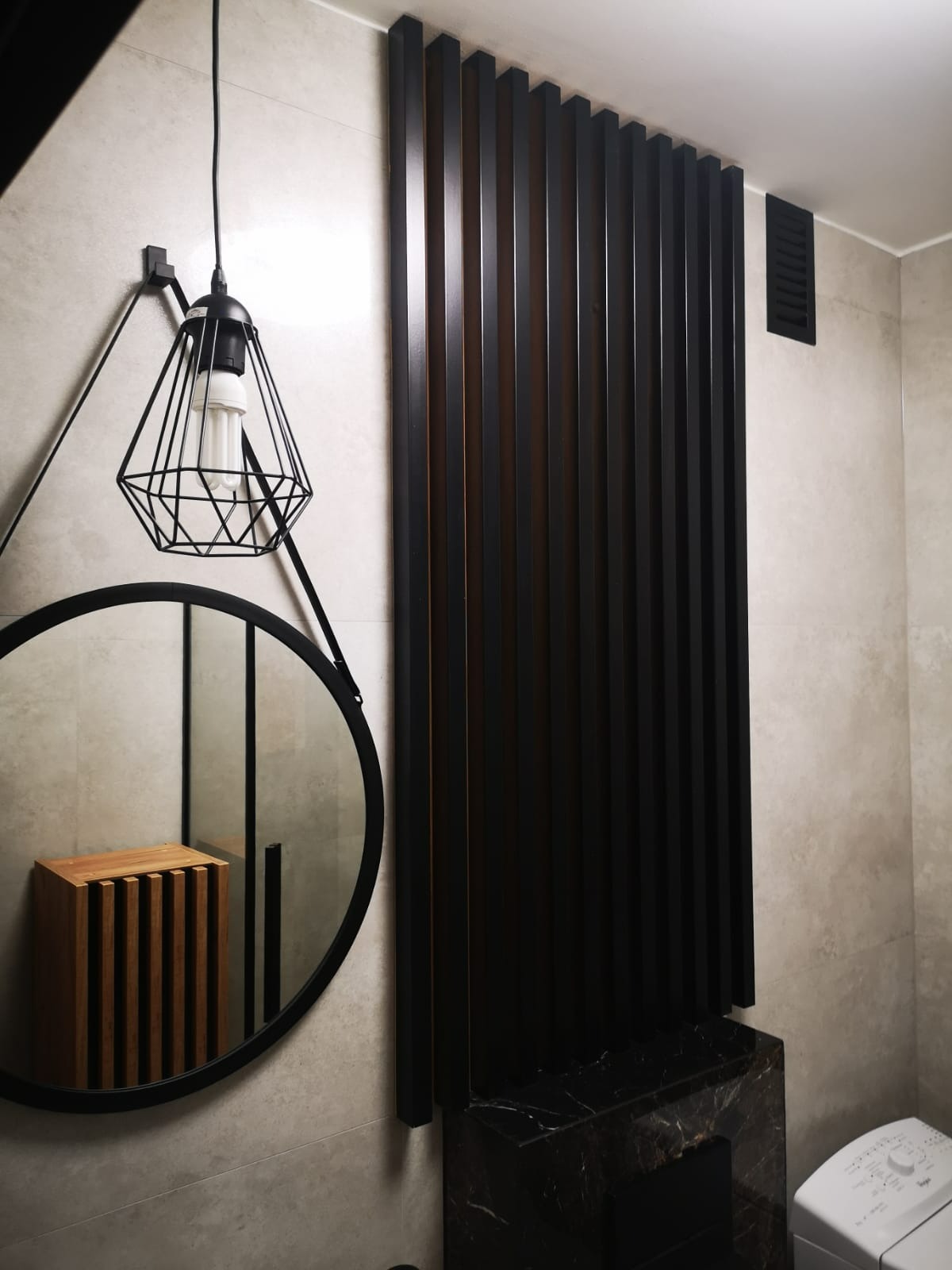 Nowoczesna łazienka z czarnymi lamelami, okrągłym lustrem i lampą w stylu industrialnym. Umywalka z czarnego marmuru i pralka w tle. Minimalistyczny design.