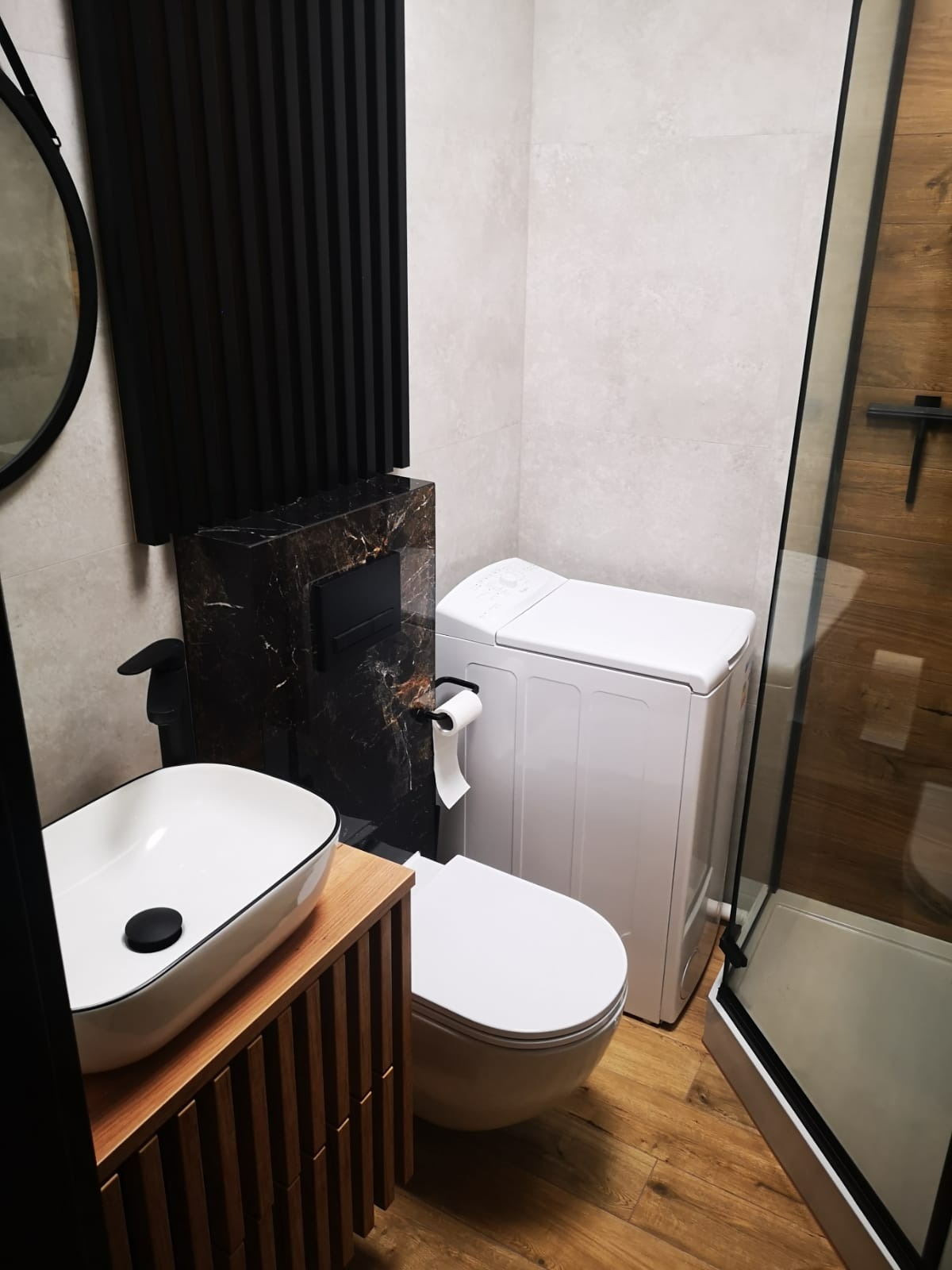 Nowoczesna łazienka z drewnianą podłogą i szafką pod umywalkę, marmurową obudową WC, białą pralką i kabiną prysznicową z drewnianymi panelami.