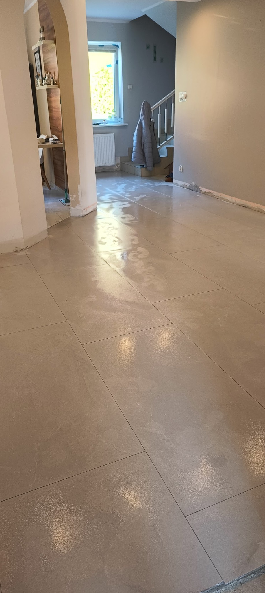 Beżowe, błyszczące płytki podłogowe ułożone w salonie. Widoczne przejście do schodów i częściowo umeblowane wnętrze. Świeżo położona podłoga.