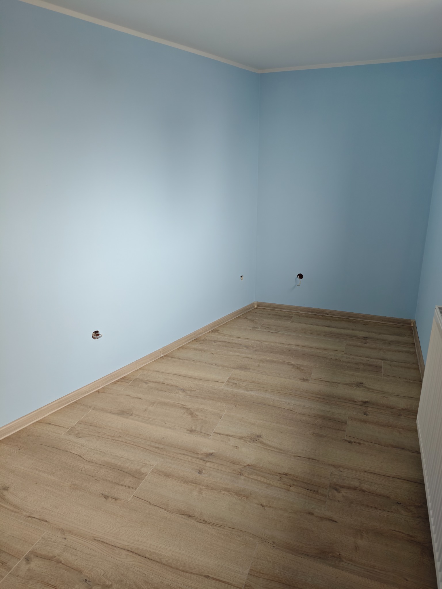 Jasne panele podłogowe ułożone w rogu pokoju z błękitnymi ścianami. Widoczne listwy przypodłogowe i otwory na gniazdka elektryczne. Grzejnik z boku kadru.