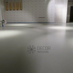 DECOR PROFESSIONAL - Jasnoszara, gładka posadzka w pomieszczeniu z białą cegłą na ścianie. Widoczne narzędzia malarskie i logo firmy Decor Professional.