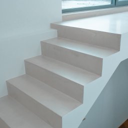 DECOR PROFESSIONAL - Minimalistyczne schody z mikrocementu w jasnym wnętrzu, z widocznym fragmentem parkietu i oknem w tle. Gładka powierzchnia, nowoczesny design.
