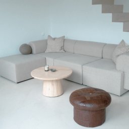 DECOR PROFESSIONAL - Jasnoszara, modułowa sofa narożna z poduszkami, okrągły stolik kawowy i pufa na jasnej posadzce. W tle minimalistyczne schody i ściana w odcieniach bieli.