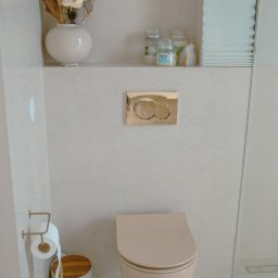 DECOR PROFESSIONAL - Beżowa toaleta podtynkowa z złotym przyciskiem spłukującym, w łazience wykończonej mikrocementem. Na półce dekoracje: suszone kwiaty i świece.