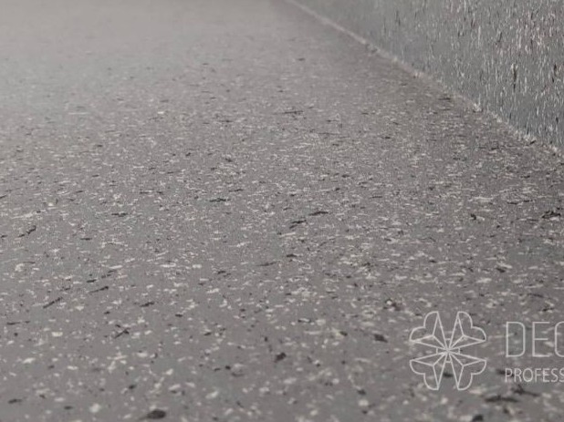 Szara posadzka żywiczna z drobnymi, ciemnymi i jasnymi inkluzjami, widoczna przy ścianie. Niska perspektywa podkreśla teksturę i gładkość powierzchni.