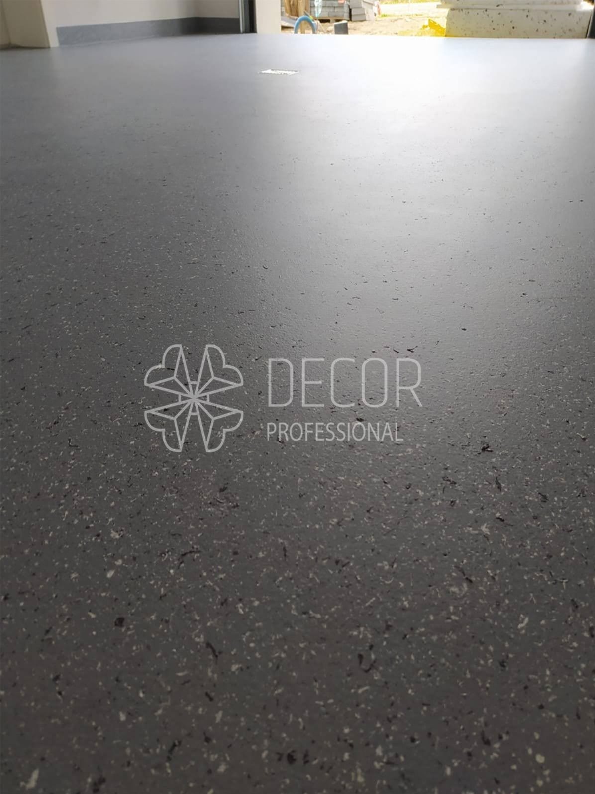 Szara, gładka posadzka z delikatnym wzorem, wykonana w technologii żywicznej. Widoczne logo firmy w dolnej części kadru. W tle fragment otwartego pomieszczenia.