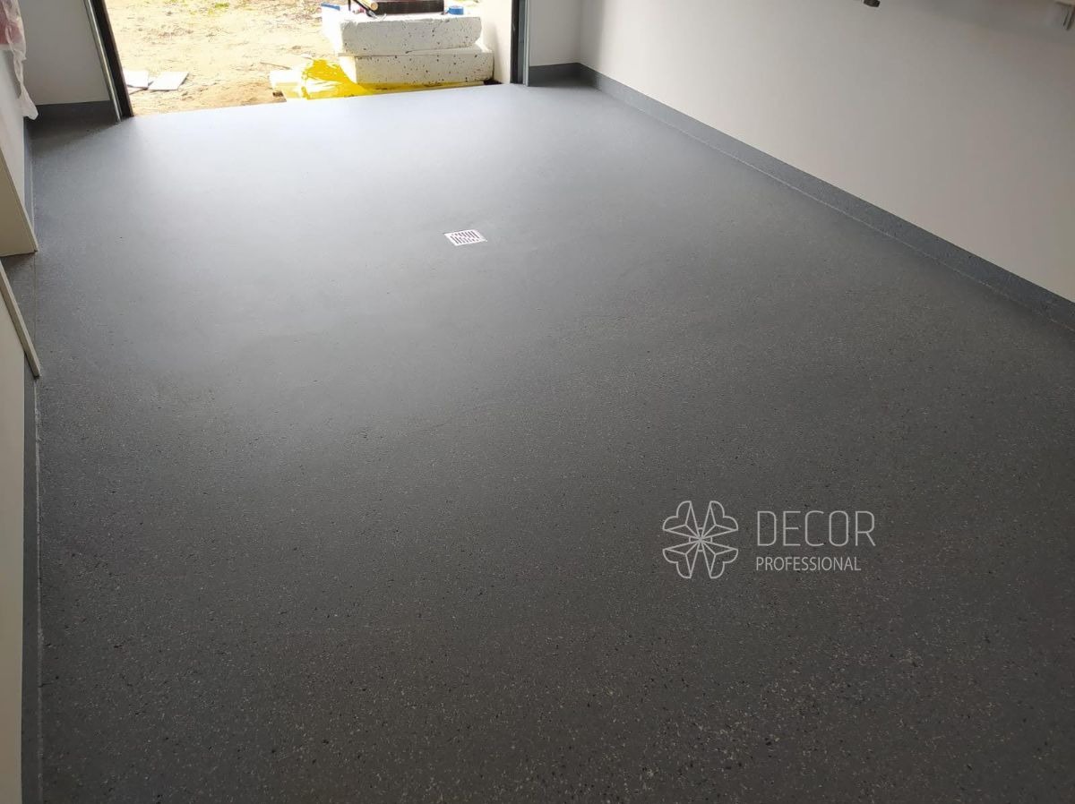 Szara posadzka żywiczna w garażu z odpływem liniowym, wykończona listwą przypodłogową. Widoczny fragment otwartej bramy garażowej i logo firmy Decor Professional.